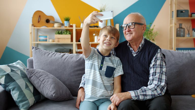 Smiling grandfather and grandson taking a selfie on a cozy living room sofa, capturing joyful family moments.